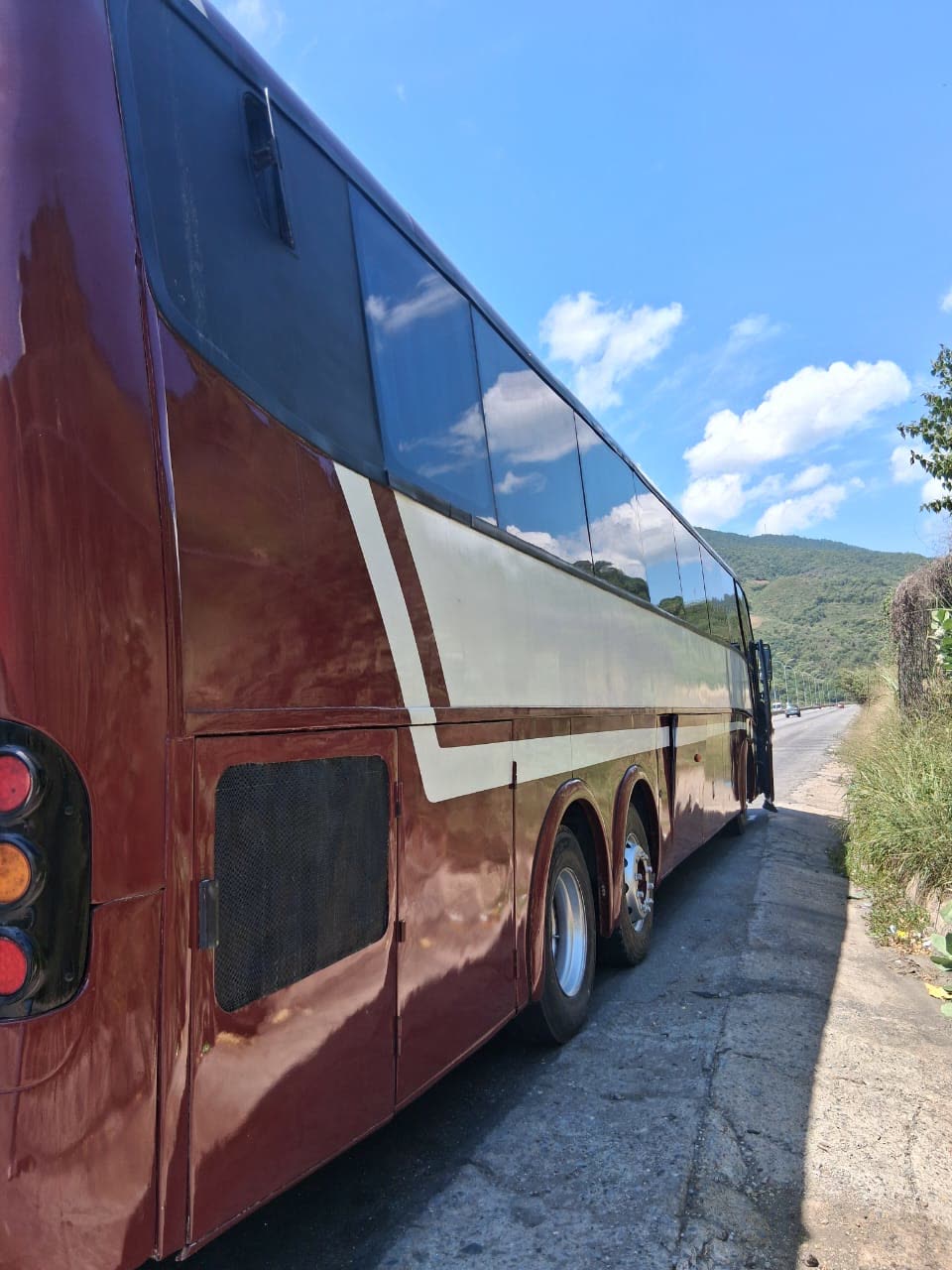 Lateral y parte trasera del autobús vinotinto con franjas blancas, estacionado en una carretera con una montaña al fondo.
